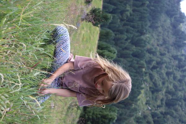 A young person with blonde hair sits amidst tall green grass on a sunny hillside, looking down intently at something in their hands